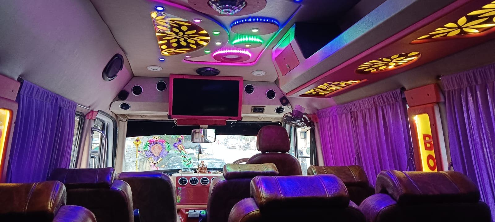 Wide view of a Tempo Traveller cabin with brown leather seats, ceiling lights and a TV